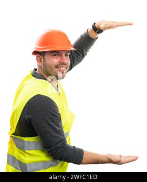 Happy builder man with trustworthy friendly expression presenting large object copyspace between hands wearing work attire helmet and vest isolated on Stock Photo