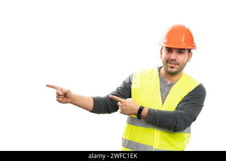 Cheerful male constructor smiling pointing at blank copyspace for advertising using index fingers wearing work attire vest and orange safety helmet is Stock Photo
