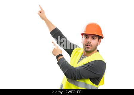 Trustworthy friendly male builder with surprised expression wearing fluorescent yellow safety vest and orange hardhat pointing index fingers at blank Stock Photo