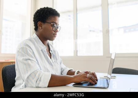 Confident serious young business woman, female freelancer, programmer, ceo corporate leader wearing smart casual shirt sitting inside and using laptop in office, typing Stock Photo