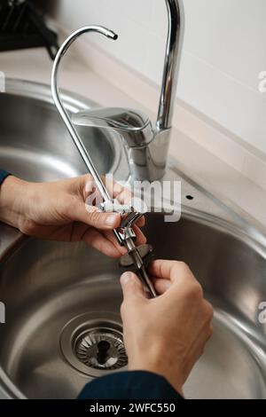 a caucasian man, in blue overalls, installs an extra faucet in a metal kitchen sink Stock Photo