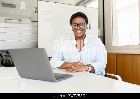 Cheerful attractive african-american female employee, manager, teamlead using laptop sitting in contemporary office at workplace, confident young multiracial businesswoman looking at camera, smiling Stock Photo