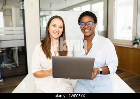 Two happy diverse female colleagues African American and Caucasian women looking at camera standing in office and holding laptop, discussing strategy together Stock Photo