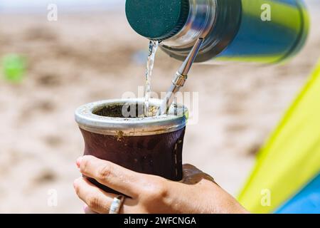 Woman serving hot water of a thermos in a Mate with yerba. Drinking Mate on the beach. Stock Photo