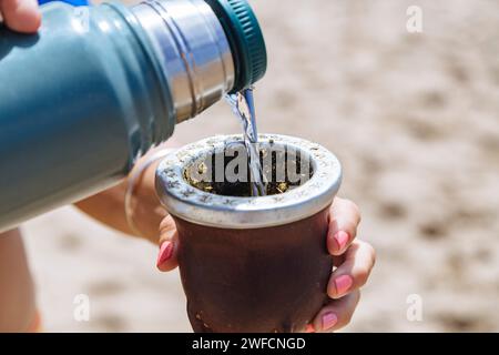 Woman serving hot water of a thermos in a Mate with yerba. Drinking Mate on the beach. Stock Photo