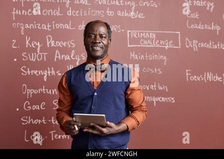 Waist up portrait of Black senior college professor wearing bowtie and smiling at camera by chalkboard copy space Stock Photo