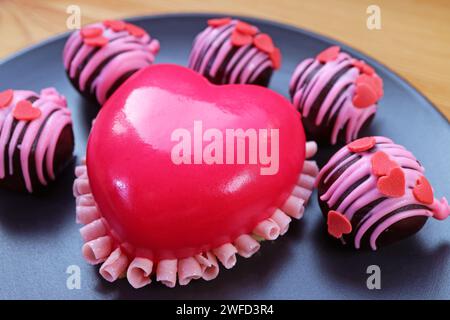 Closeup of Vivid Pink Heart Shaped Strawberry Mousse Cake with Chocolate Cake Balls on Black Plate Stock Photo