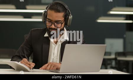Focused Arabian male Indian businessman wear headphones ethnic business man studying working in office with laptop writing notes write in notebook Stock Photo