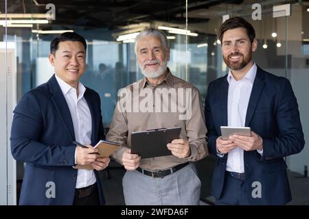 Three diverse professional businessmen smiling and standing together in an office setting, showcasing teamwork and collaboration. Stock Photo