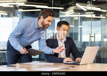 Two professional male colleagues collaborate at a work desk in a bright modern office, discussing business strategies on a laptop. Stock Photo