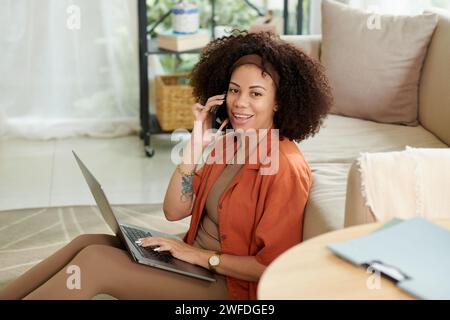 Portrait of cheerful woman answering phone call from colleague when working on laptop at home Stock Photo
