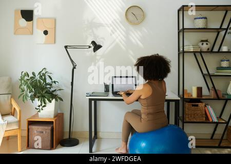 Woman sitting on fitness ball when working on laptop at home, view from behind Stock Photo