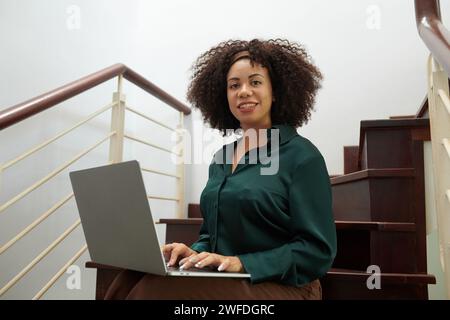 Portrait of cheerful Latin businesswoman sitting on steps and working on laptop Stock Photo