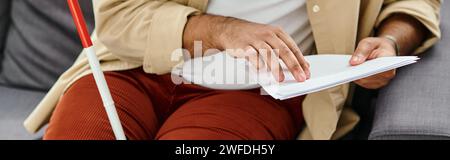 cropped view of indian blind man in comfy clothing sitting on sofa and reading braille code, banner Stock Photo