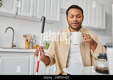 attractive indian man with visual impairment sitting and drinking tasty coffee while on kitchen Stock Photo