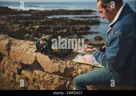 Happy man tourist adventurer in casual denim, using compass and binoculars, reading navigational map while sitting on the rock by sea. People. Lifesty Stock Photo