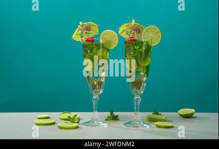 Two cocktails in long glass  filled with ice and lemon slices garnished with small yellow umbrellas, cherries and lemon slices on a white table agains Stock Photo