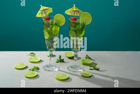 Two cocktails in long glass filled with ice and lemon slices garnished with small yellow umbrellas, cherries and lemon slices on a white table against Stock Photo