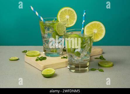 Two cocktails in glass glasses filled with ice and lemon slices garnished with lemon slices and sorbets on a wooden board against turquoise background Stock Photo