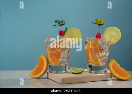 Two cocktails in glass glass filled with ice and orange slice garnished with lemon slice, cherry and butterfly pin on wooden board with pieces of oran Stock Photo