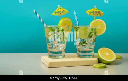 Two cocktails in glass  filled with ice and lemon slices garnished with lemon slices, yellow umbrellas and sorbets on a wooden board against turquoise Stock Photo