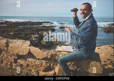Smiling young male adventurer tourist traveler sitting on a rock by sea near his backpack, holding binoculars and checking directions using maps and c Stock Photo