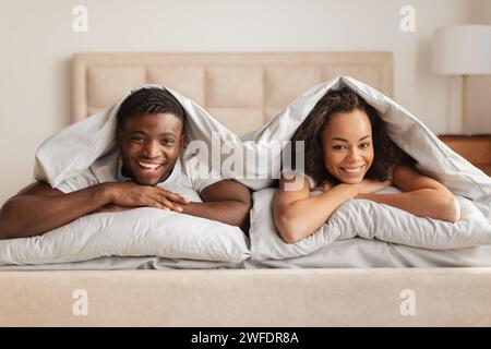 Smiling African American Young Spouses Lying Under Blanket In Bed Stock Photo