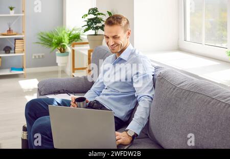 Happy man sitting on the sofa, watching a video on laptop and drinking hot tea from a thermos Stock Photo