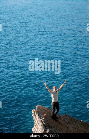 Smiling young man stands on a stone ledge above the sea with his hands up Stock Photo