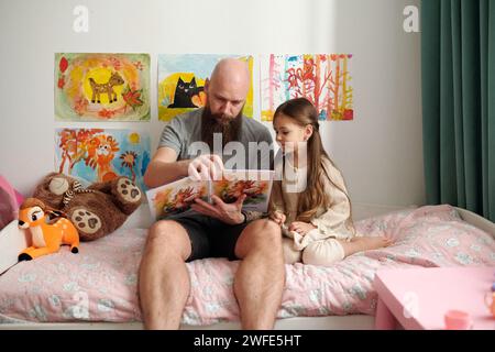 Bearded man with open book of comics with pictures sitting on bed next to his adorable little daughter and reading her new stories Stock Photo