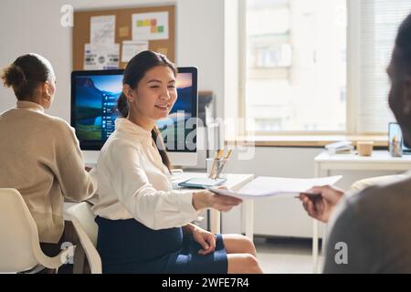 Happy young Asian businesswoman taking documents from hand of African American male colleague while sitting next to coworker Stock Photo