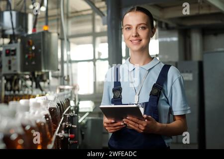 Young smiling female inspector with tablet looking at camera while standing by assembly line with row of capped bottles Stock Photo