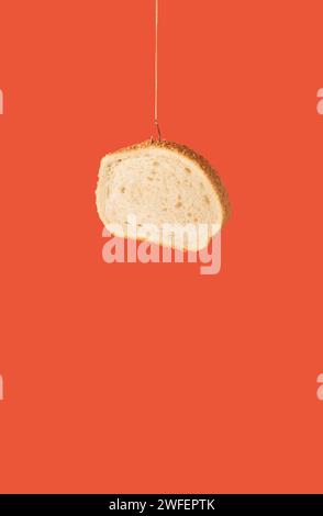 Slice of bread hanging on fish hook against orange background. Minimal food composition. Stock Photo