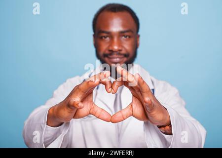 Close-up of a heart shape made by fingers of an African american medical practitioner. Black male professional doctor dressed in lab coat doing love hand gestures toward the camera. Stock Photo