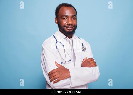 Portrait of black male physician confidently posing with crossed arms for the camera in studio. African American medical specialist wearing lab coat is standing against isolated backdrop. Stock Photo