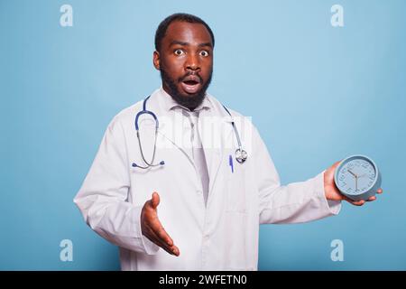 Medical practitioner looking surprised running out of time holding alarm clock while wearing lab coat and stethoscope. African american doctor panicking for being late for an appointment. Stock Photo