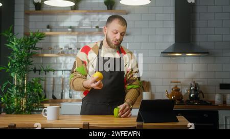 Vlogger male in an apron talking into tablet computer, recording video cooking lesson about vegetarian meal or salad in modern kitchen at home. Online Stock Photo