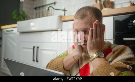 Shocked man sit on the floor in kitchen typing on laptop enjoying surfing internet, using social media, chatting with friends Stock Photo