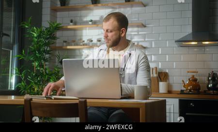 Experienced doctor wearing white coat using laptop computer studying patients reports at home office. Male practitioner doing paperwork Stock Photo