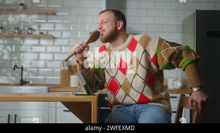 Cozy man singing into a ladle like a microphone while sit in the kitchen at home Stock Photo