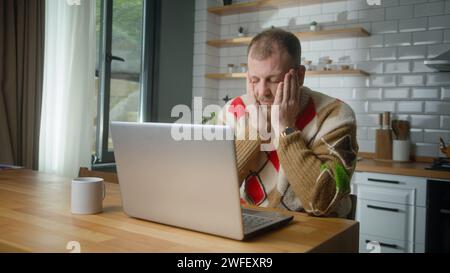man sit in the kitchen experiences severe pain in neck, rubbing it to relieve muscle tension, massaging his neck and taking a break from working on la Stock Photo