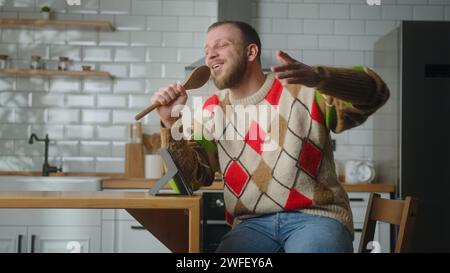 Happy adult man singing into a ladle like a microphone while sit in the kitchen at home Stock Photo