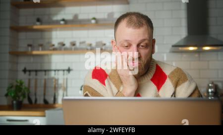adult man sit in kitchen surprised, stunned while watching movie or video or reading news on laptop at home Stock Photo