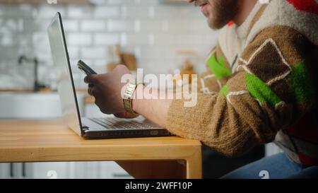Man sit in kitchen with laptop, surfing internet on mobile phone, reading media news, scrolling social networks, using mobile applications on smartpho Stock Photo