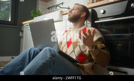 Man sitting on the floor in kitchen using laptop having anxiety and stress. Upset man reacting to loss, bad news Stock Photo