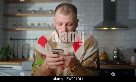 Smiling man sit in kitchen surfing internet on mobile phone, checking email, reading media news, scrolling social networks, using mobile applications Stock Photo