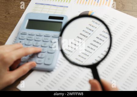 Woman looking at accounting document through magnifying glass while using calculator at wooden table, closeup Stock Photo