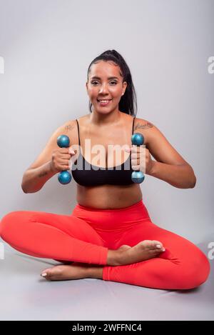 Beautiful, brunette, happy and smiling woman in gym clothes, sitting holding dumbbells. Healthy life. Against gray background. Stock Photo