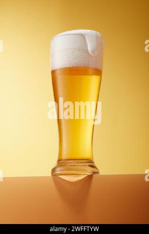 A transparent glass filled with beer, placed on podium against a gradient golden background. The frothy foam on top of beer. Alcoholic beverages and d Stock Photo