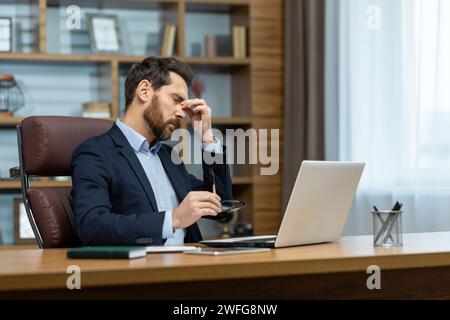 Overworked bearded male holding eyeglasses and rubbing dry irritated eyes after looking at laptop screen too long. Exhausted man having bad vision and making break for sight distraction from work. Stock Photo